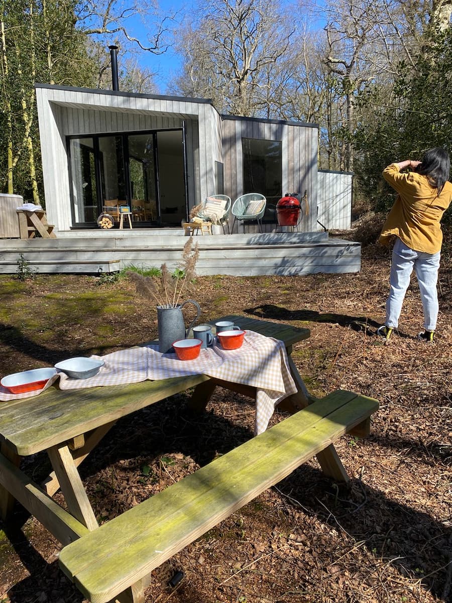 Behind the scenes photo shoot. In the foreground is a picnic table styled with tablecoth and bowls, in the background a photographer is taking photos of the koto cabin situated within woodland