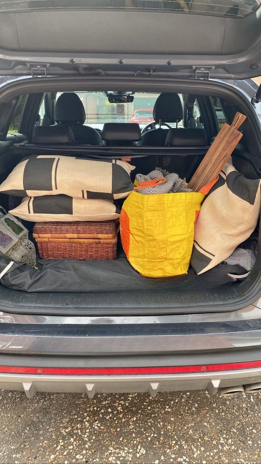Car boot filled with props for a photo shoot Hessian and black cushions on the top of a picnic basket to the left, an orange Sainsbury's bag for life filled with a grey throw and wooden serving board