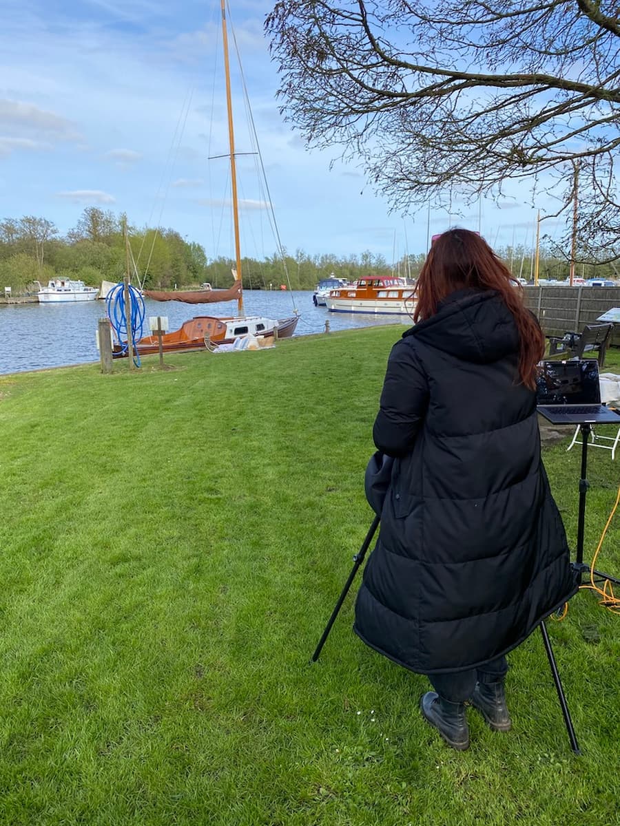 Woman dressed in a long black puffa coat positioned to the right setting up for a photo shoot of a sail boat which is in the background on the Norfolk