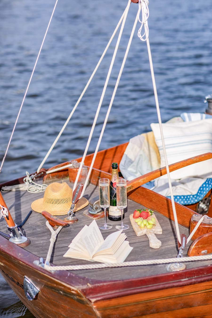 Styled shot at the front of a sail boat consisting of a straw hat, champagne flutes, an open book and a wooden board with grapes and strawberries The sun is glowing perfectly providing the all important golden glow