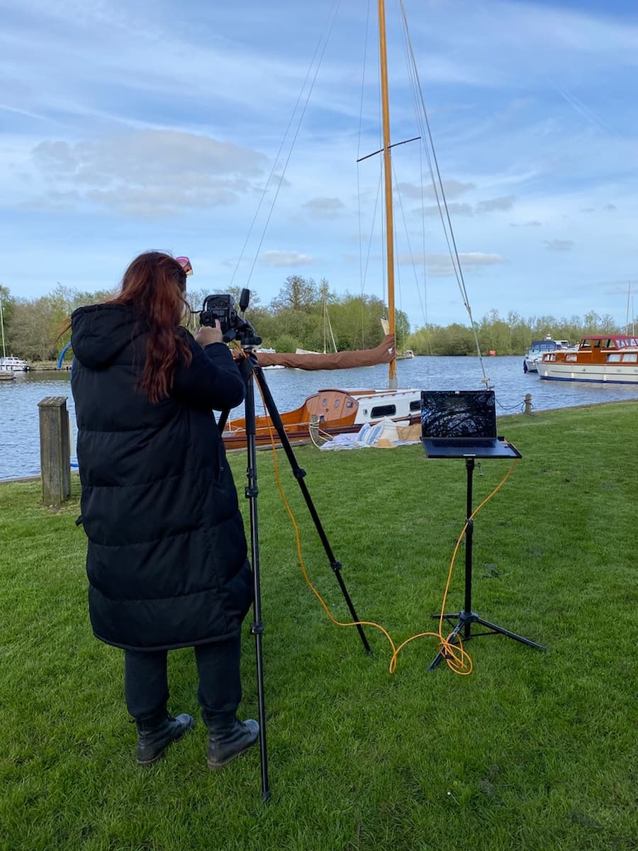 Woman in long black puffa coat positioned to the left in front of a tripod, looking through a camera which is connected to a laptop for a photo shoot at the Norfolk broads. A sail boat is in the background on the water.