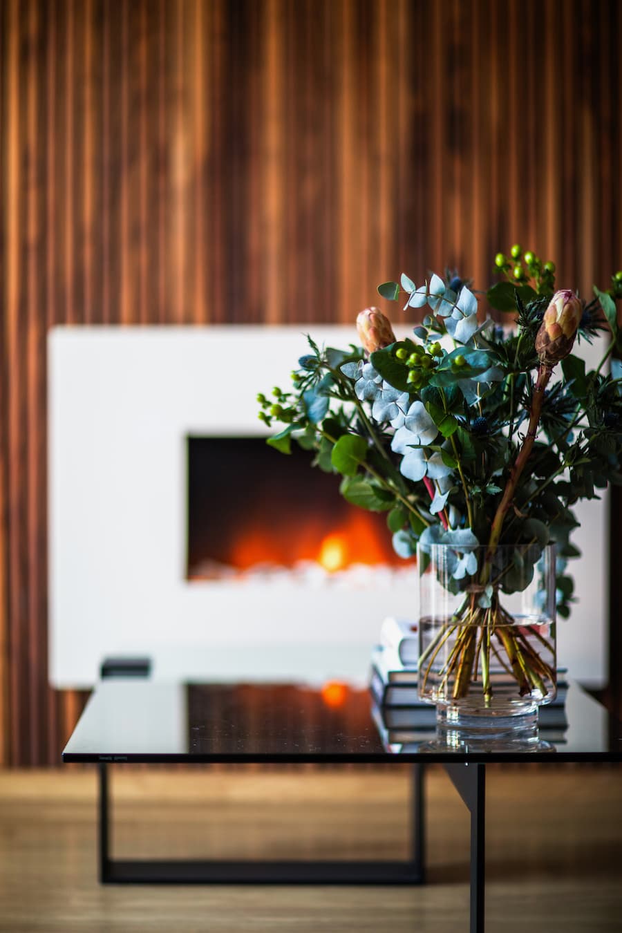 minimalist interior decor with glass coffee table and a simple bouquet of flowers in a glass vase postioned in front of a fire and wood wall panelling