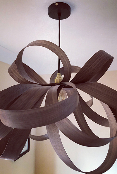 A modern chandelier hanging from a white ceiling. The chandelier is made of dark wood and has a unique, abstract design with curved, overlapping pieces. The light bulb is visible in the centre of the chandelier and is turned on, casting a warm glow.