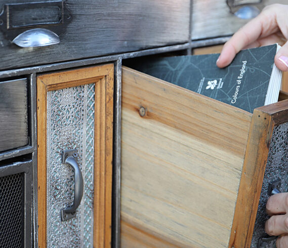 A hand pulling out a drawer from a wooden cabinet. The cabinet has multiple drawers with different designs and textures. The drawer being pulled out is made of wood and has a colour chart with a dark grey cover being pulled out.