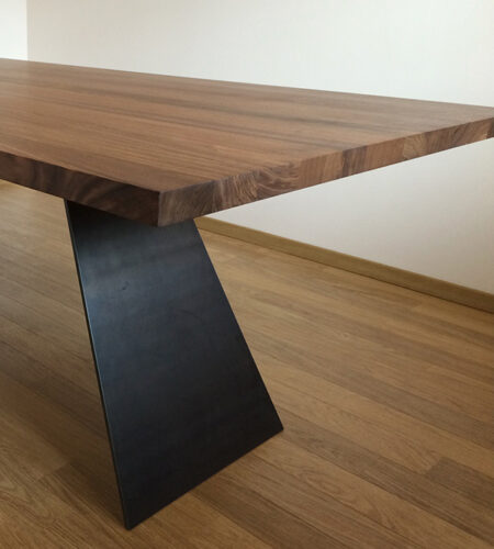 Cropped version of a wooden table with a walnut finish. The table has a rectangular top with a live edge on the long sides. The table is supported by two triangular legs that are in steel. The table is in a room with a wooden flooring and white walls. The image is taken from a low angle, slightly to the side of the table.