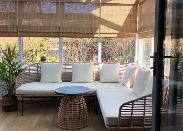 A sunroom with a modern design. The sunroom has a wooden floor and a glass roof. The furniture consists of a white corner sofa with white cushions, a round coffee table with a rattan base. There is a potted plant in the corner of the room. The background shows a garden with trees and shrubs. The room is bright and airy with natural light coming in from the glass roof and windows
