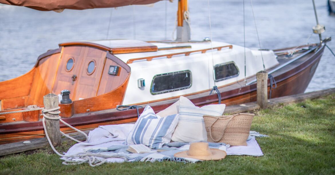 Sailboat on the Norfolk broads moored up on an embankment with a blanket and selection of blue and white striped cushions, a straw hat and bag